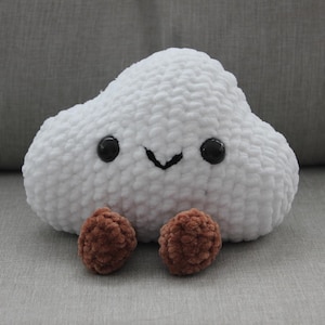 May include: A handmade white cloud-shaped plush toy with black button eyes and a stitched smile. Two brown, round, plush shapes are attached at the bottom, resembling feet. The toy is made of soft, textured material.