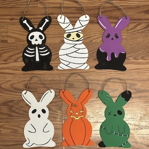 Può includere: Sei decorazioni di Halloween a forma di coniglietto in legno. Ogni coniglietto è dipinto con un design spettrale diverso: uno scheletro, una mummia, una melma viola, un fantasma, una zucca e un mostro di Frankenstein. Ognuno ha un anello di spago per appenderlo.
