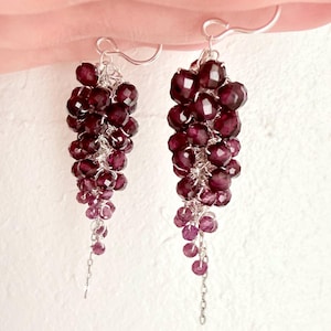 May include: A pair of dangle earrings featuring clusters of faceted, deep burgundy garnet beads wired onto silver-toned metal. The beads are arranged in a cascading, grape-like formation that tapers toward the bottom, suspended from simple silver hooks.