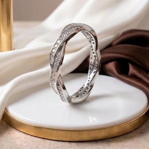 Può includere: Un anello color argento caratterizzato da un design a fascia intrecciata e ritorta. La superficie della fascia è incastonata con numerose piccole pietre trasparenti a taglio rotondo che catturano la luce, creando un effetto scintillante continuo.