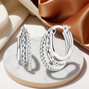 Könnte beinhalten: Zwei silberne Creolen mit dreilagigem Design. Ein Ohrring hat eine Reihe kleiner, klarer Steine. Die Ohrringe werden auf einer weißen und goldenen Oberfläche mit einem weichen, neutralen Hintergrund präsentiert.