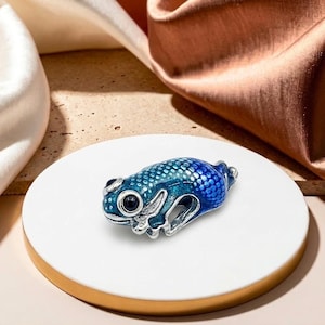 May include: A blue and silver frog charm. The charm features a detailed design with a speckled body and large black eyes. The frog is displayed on a white circular surface.