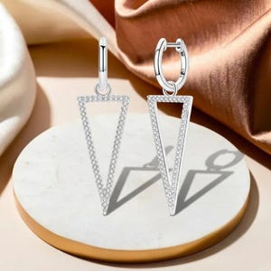 Puede incluir: Un par de pendientes de aro plateados con un diseño triangular abierto colgante. El triángulo está delineado con pequeñas piedras claras y brillantes. Los pendientes se exhiben sobre una superficie blanca y dorada contra un fondo suave y neutro.