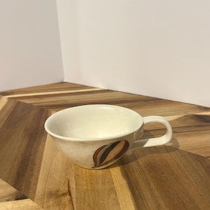 May include: Cream-colored teacup with a curved handle and a decorative design of red, brown, and green shapes. The cup sits on a wooden surface, showcasing its unique design and shape.