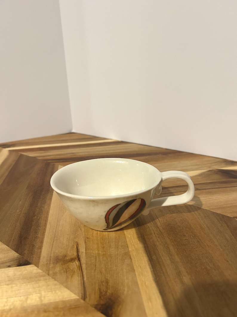 May include: Cream-colored teacup with a curved handle and a decorative design of red, brown, and green shapes. The cup sits on a wooden surface, showcasing its unique design and shape.