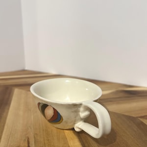 May include: Cream-colored ceramic teacup with a rounded handle. The cup features a colorful abstract design on the side, with shades of brown, red, blue, and yellow. The cup is set on a wooden surface.