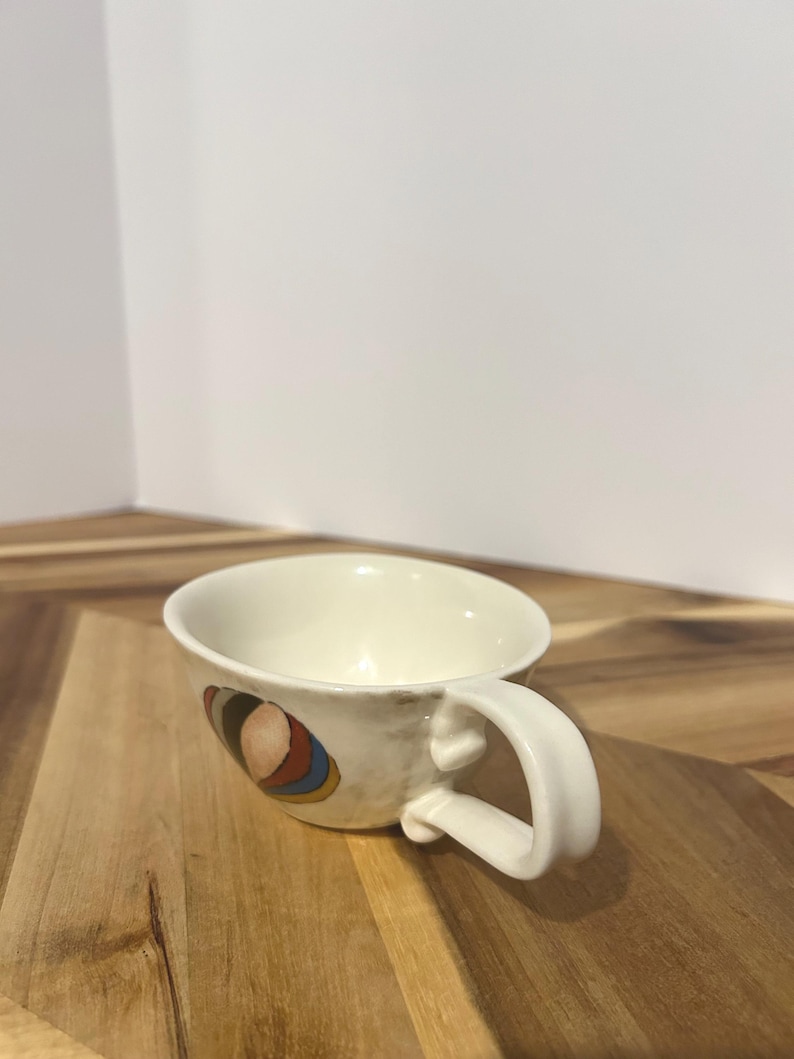 May include: Cream-colored ceramic teacup with a rounded handle. The cup features a colorful abstract design on the side, with shades of brown, red, blue, and yellow. The cup is set on a wooden surface.