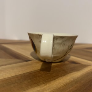 May include: A vintage teacup with a cream-colored handle and a floral design. The cup has a beige background with brown and yellow floral accents. The cup is sitting on a wooden surface.