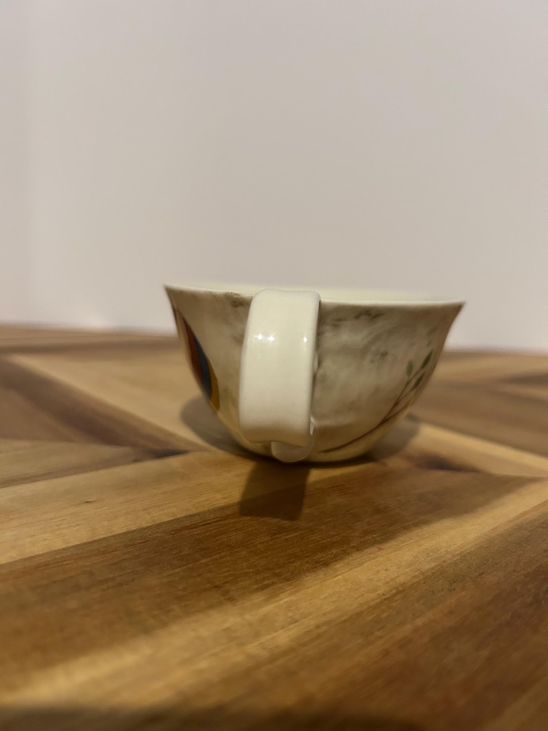 May include: A vintage teacup with a cream-colored handle and a floral design. The cup has a beige background with brown and yellow floral accents. The cup is sitting on a wooden surface.