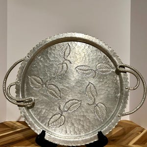 May include: A silver-colored, round serving tray with a hammered texture and embossed leaf design. The tray has two curved handles and a scalloped edge. The tray is displayed on a black stand.