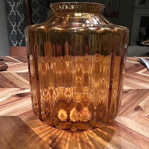 May include: A large, amber-colored glass vase with a ribbed design. The vase has a wide, cylindrical body and a slightly flared neck. The glass reflects light, creating a warm, inviting glow. The vase sits on a wooden table.