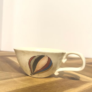 May include: Cream-colored ceramic teacup with a curved handle. The cup features a colorful hot air balloon design in shades of red, blue, orange, and black. The cup sits on a wooden surface.