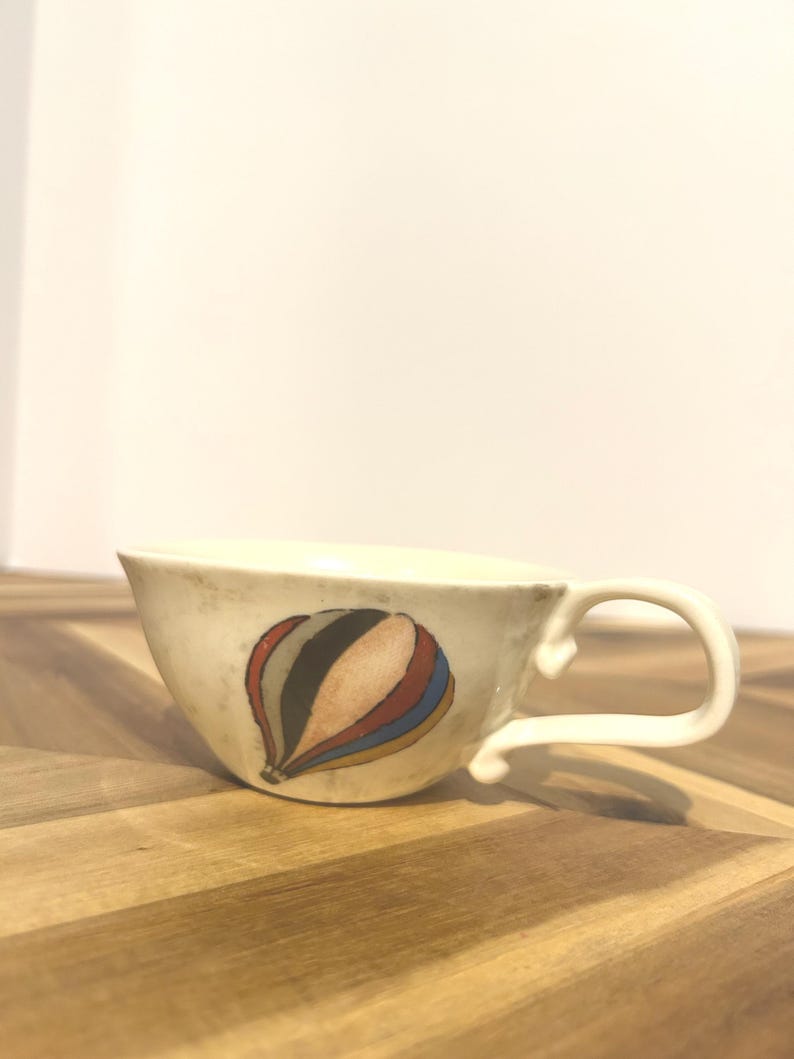 May include: Cream-colored ceramic teacup with a curved handle. The cup features a colorful hot air balloon design in shades of red, blue, orange, and black. The cup sits on a wooden surface.