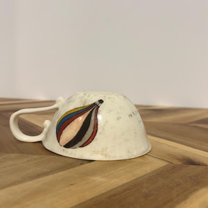 May include: Cream-colored ceramic teacup with a curved handle. The cup's exterior features a colorful abstract design with red, blue, yellow, and black curved shapes. The cup is resting upside down on a wooden surface.