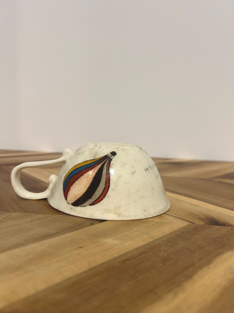 May include: Cream-colored ceramic teacup with a curved handle. The cup's exterior features a colorful abstract design with red, blue, yellow, and black curved shapes. The cup is resting upside down on a wooden surface.
