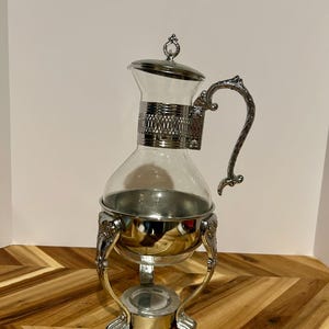 May include: A vintage silver-plated coffee server with a clear glass carafe. The server has an ornate handle and decorative base with claw feet. The lid is silver with a small decorative finial. The server is designed to keep coffee warm.