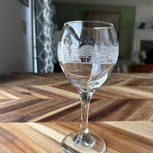 May include: A clear wine glass with a detailed etched winter scene design. The glass features a slender stem and a round base. The etched design includes trees and a snowy landscape. The glass is sitting on a wooden table.
