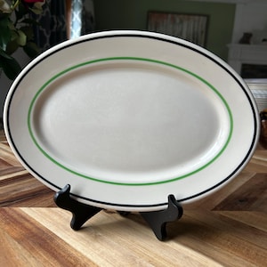 May include: An oval, off-white serving platter with a green and black border. The platter is displayed on a black stand, showcasing its design. The plate is made of ceramic and is suitable for serving food.