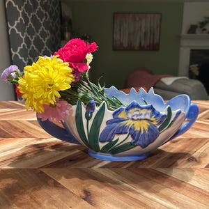 May include: A ceramic floral centerpiece with a blue rim and handles, featuring a painted iris design. The vase holds a vibrant arrangement of yellow, pink, and red flowers, set on a wooden table.