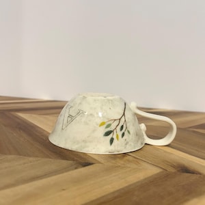 May include: A white ceramic teacup with a handle, decorated with a branch design and the letter "A". The branch has green and yellow leaves. The cup is resting on a wooden surface.
