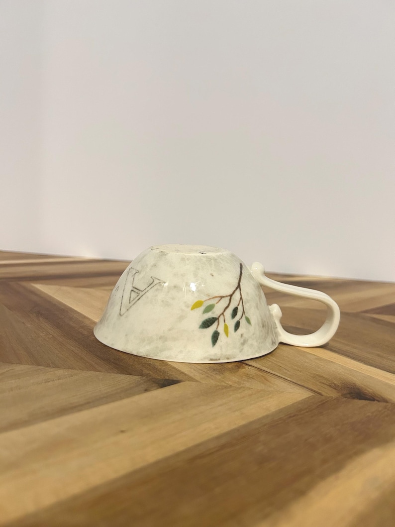 May include: A white ceramic teacup with a handle, decorated with a branch design and the letter "A". The branch has green and yellow leaves. The cup is resting on a wooden surface.