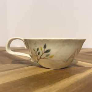 May include: Cream-colored teacup with a curved handle. The cup features a painted branch with green and yellow leaves and the letter "A". The cup is set on a wooden surface.