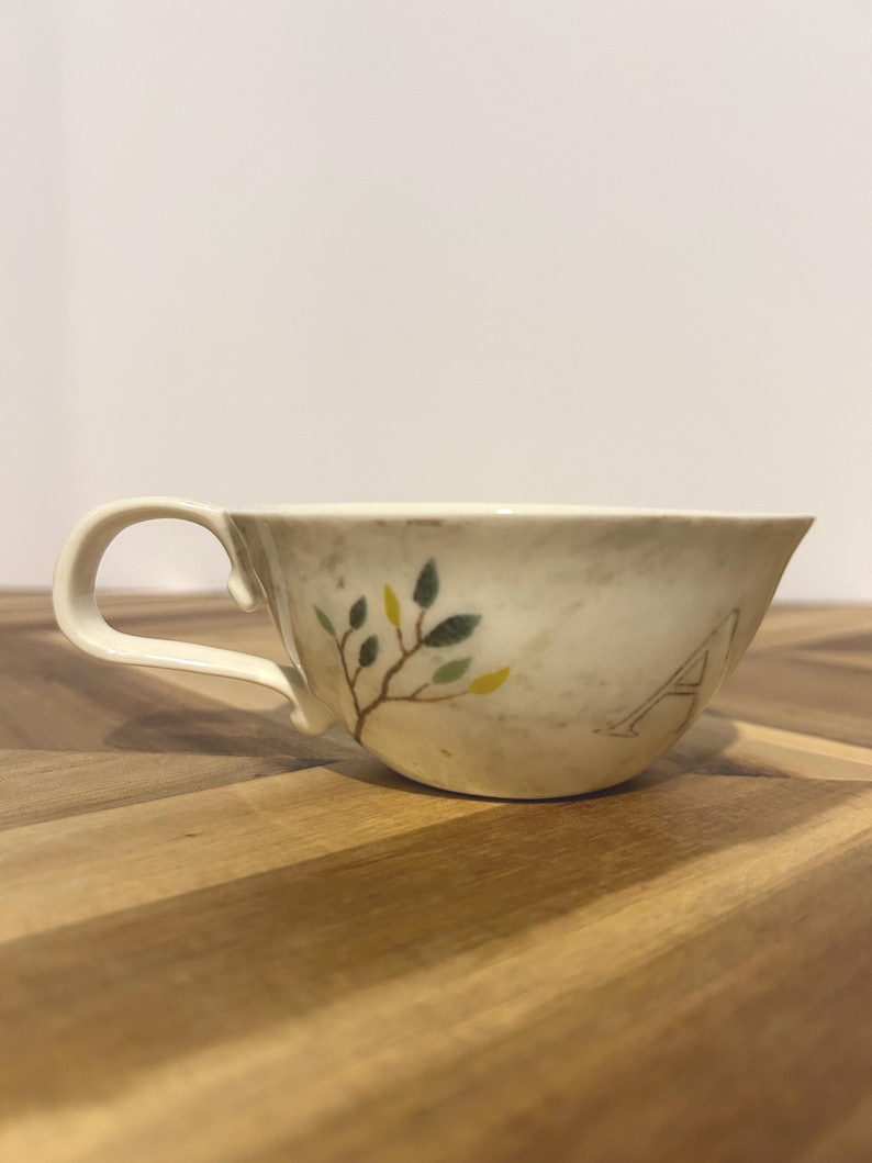 May include: Cream-colored teacup with a curved handle. The cup features a painted branch with green and yellow leaves and the letter "A". The cup is set on a wooden surface.