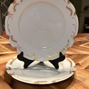May include: Two white porcelain plates with gold trim and scalloped edges. The plates are displayed on a black stand, showcasing their elegant design. The gold detailing adds a touch of sophistication to the tableware.