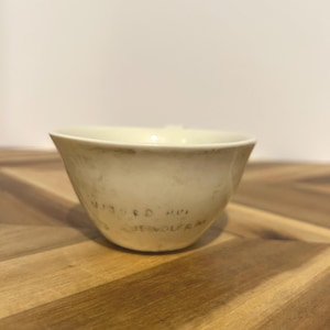 May include: A cream-colored ceramic bowl with a slightly flared rim. The bowl has a matte finish and appears to be handmade. The text "WIEBKE HVI DE WOLFRA" is written on the side of the bowl in a dark color.