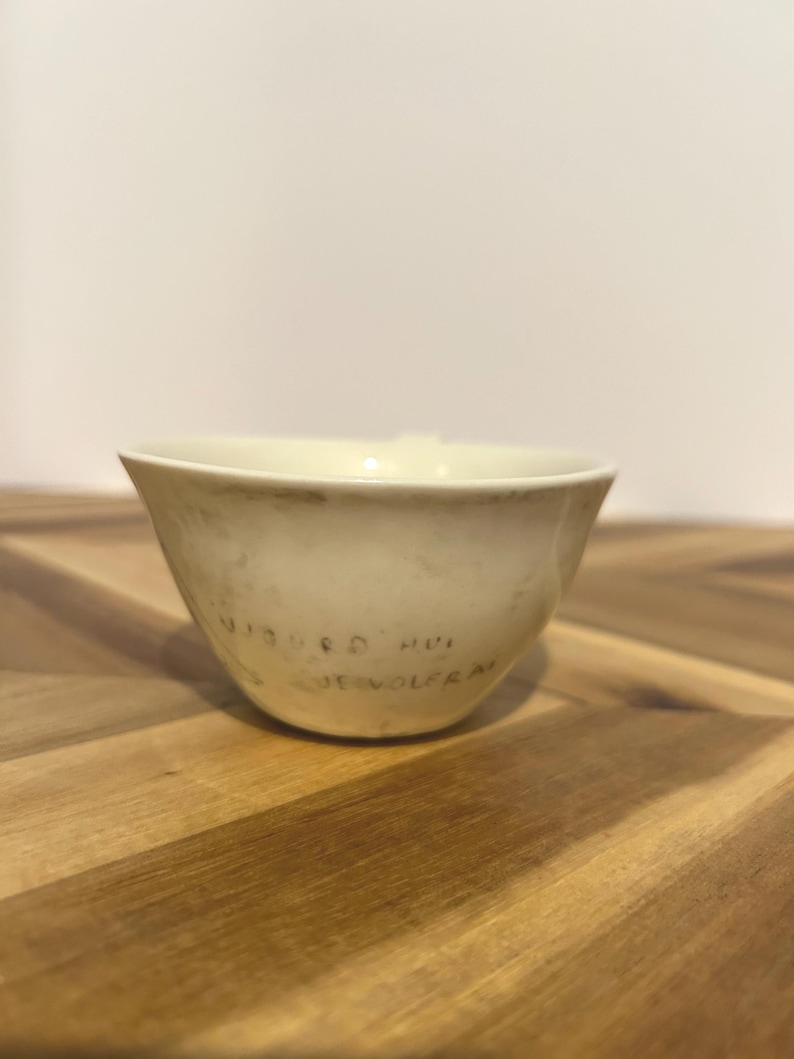 May include: A cream-colored ceramic bowl with a slightly flared rim. The bowl has a matte finish and appears to be handmade. The text "WIEBKE HVI DE WOLFRA" is written on the side of the bowl in a dark color.