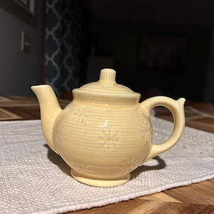 May include: A small, pale yellow teapot with a lid and handle. The teapot has a rounded body with embossed snowflake designs and sits on a small base. The spout and handle curve gracefully. The teapot is on a white textured placemat on a wooden table.