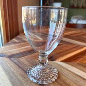 May include: A clear, vintage-style stemmed glass with a textured base and a swirling design in the bowl. The glass sits on a wooden surface, reflecting light and creating a sense of elegance. Ideal for serving beverages.