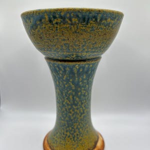 May include: A ceramic pedestal bowl with a textured surface. The bowl is teal and brown with a speckled design. The base is a flared shape with a brown rim. The bowl is likely used for decorative purposes.