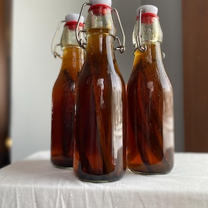 May include: Three glass bottles filled with a dark amber liquid and vanilla beans. Each bottle has a white stopper with a red ring and a metal clasp. The bottles are arranged on a white surface.