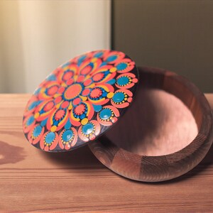 May include: A wooden box with a decorative lid. The lid features a colorful mandala design with orange, blue, and yellow accents. The box is open, revealing a light-colored interior. The box sits on a wooden surface.