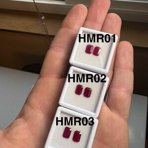 May include: Three white display boxes, each containing two rectangular, deep red gemstones. The boxes are labeled HMR01, HMR02, and HMR03 in black text. The gemstones are displayed against a white background.