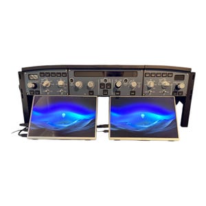 May include: A black and gray aircraft cockpit simulator with multiple dials, switches, and screens. Two laptops with blue and white screens are positioned below the cockpit panel. The simulator is designed for flight training or entertainment.