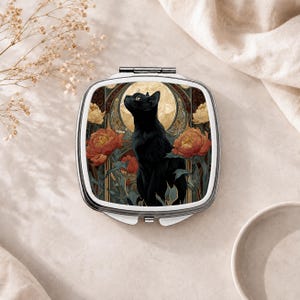 May include: A square metal compact mirror featuring a black cat illustration centered against a golden circular background. The design includes orange and yellow peony flowers with green leaves, framed by dark decorative patterns on a silver-toned case.