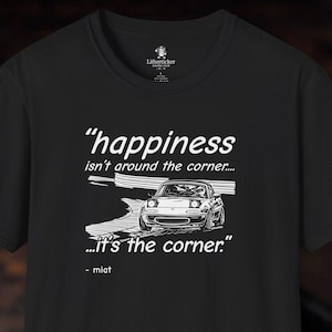 Puede incluir: Camiseta negra con un gráfico blanco de un coche y el texto "happiness isn't around the corner... ...it's the corner." La palabra "miat" está debajo del gráfico.