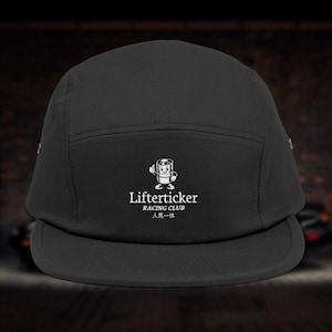Puede incluir: Gorra negra de cinco paneles con visera plana. La gorra presenta el texto blanco "Lifterticker RACING CLUB" y un gráfico de dibujos animados de un personaje sonriente. El texto japonés "人馬一体" está debajo.
