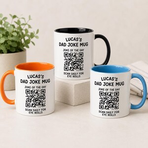 May include: Three white ceramic mugs featuring the text LUCAS'S DAD JOKE MUG, JOKE OF THE DAY, a QR code, and SCAN DAILY FOR EYE ROLLS. The mugs have black, orange, and blue handles and interior rims, respectively, and are arranged on a surface.