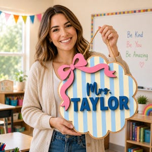 May include: A cloud-shaped wooden wall sign featuring vertical light blue and pale yellow stripes. The sign displays the text Mrs. TAYLOR in dark blue 3D lettering and is decorated with a large pink wooden bow. It hangs from a thin brown rope.