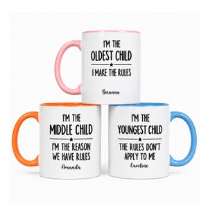 May include: Three white ceramic mugs with colored handles and rims. The top mug has a pink handle and reads I'M THE OLDEST CHILD I MAKE THE RULES Brianna. The bottom left has an orange handle and reads I'M THE MIDDLE CHILD I'M THE REASON WE HAVE RULES Amanda. The bottom right has a blue handle and reads I'M THE YOUNGEST CHILD THE RULES DON'T APPLY TO ME Caroline.