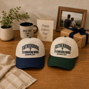 May include: Two baseball caps with the text "FATHERHOOD IS KINGDOM WORK Proverbs 22:6" in black on a cream-colored crown and a navy blue and green brim. A mug with "BEST DAD EVER" and a Father's Day card are also present.