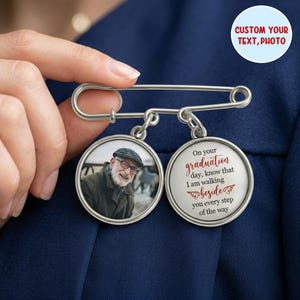 May include: A silver-tone pin with two circular pendants. One pendant features a photo of a man, the other has the text "On your graduation day, know that I am walking beside you every step of the way." The pin is held against a navy blue fabric.