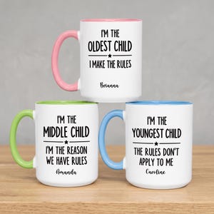 May include: Three white ceramic mugs with colored handles and rims. The top mug has a pink handle and reads I'M THE OLDEST CHILD I MAKE THE RULES Brianna. The bottom left has a green handle and reads I'M THE MIDDLE CHILD I'M THE REASON WE HAVE RULES Amanda. The bottom right has a blue handle and reads I'M THE YOUNGEST CHILD THE RULES DON'T APPLY TO ME Caroline.