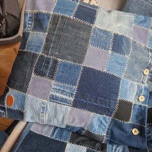 Könnte beinhalten: Ein Patchwork-Kissen aus Denim mit einer Vielzahl von Blautönen, von hell bis dunkel. Die Quadrate sind mit weißem Faden zusammengenäht. Das Kissen hat ein braunes Lederetikett und Holzknöpfe.