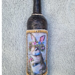 Könnte beinhalten: Eine dekorative Weinflasche mit schwarzem, strukturiertem Äußeren. Die Flasche zeigt ein detailliertes Bild eines Hasen mit Blumenkranz. Das Bild ist mit einem goldenen Rahmen versehen. Die Flasche ist mit einem schwarzen Verschluss versehen.