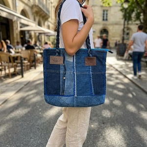 Op de afbeelding: Een rechthoekige draagtas gemaakt van verschillende tinten blauwe denim stof. De tas is voorzien van gevlochten denim schouderbanden, een felblauwe ritssluiting en twee bruine, leerachtige rechthoekige labels met de tekst AGRIES.
