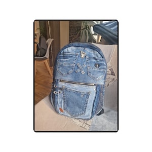 Könnte beinhalten: Ein Jeans-Rucksack mit mehreren Taschen und einem Reißverschluss. Der Rucksack besteht aus verschiedenen Blautönen, mit dekorativen Metallakzenten und einem kleinen braunen Aufnäher.